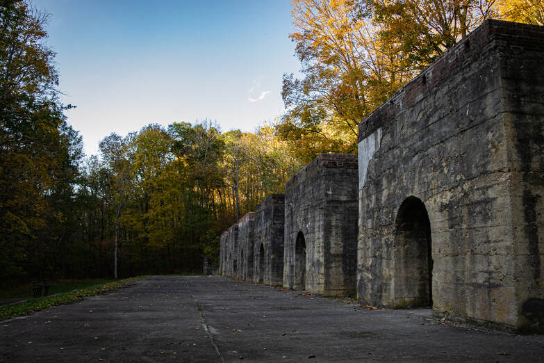 Photograph of 6 old stone archways receding in distance on cracked concrete, with sunlit fall forest behind